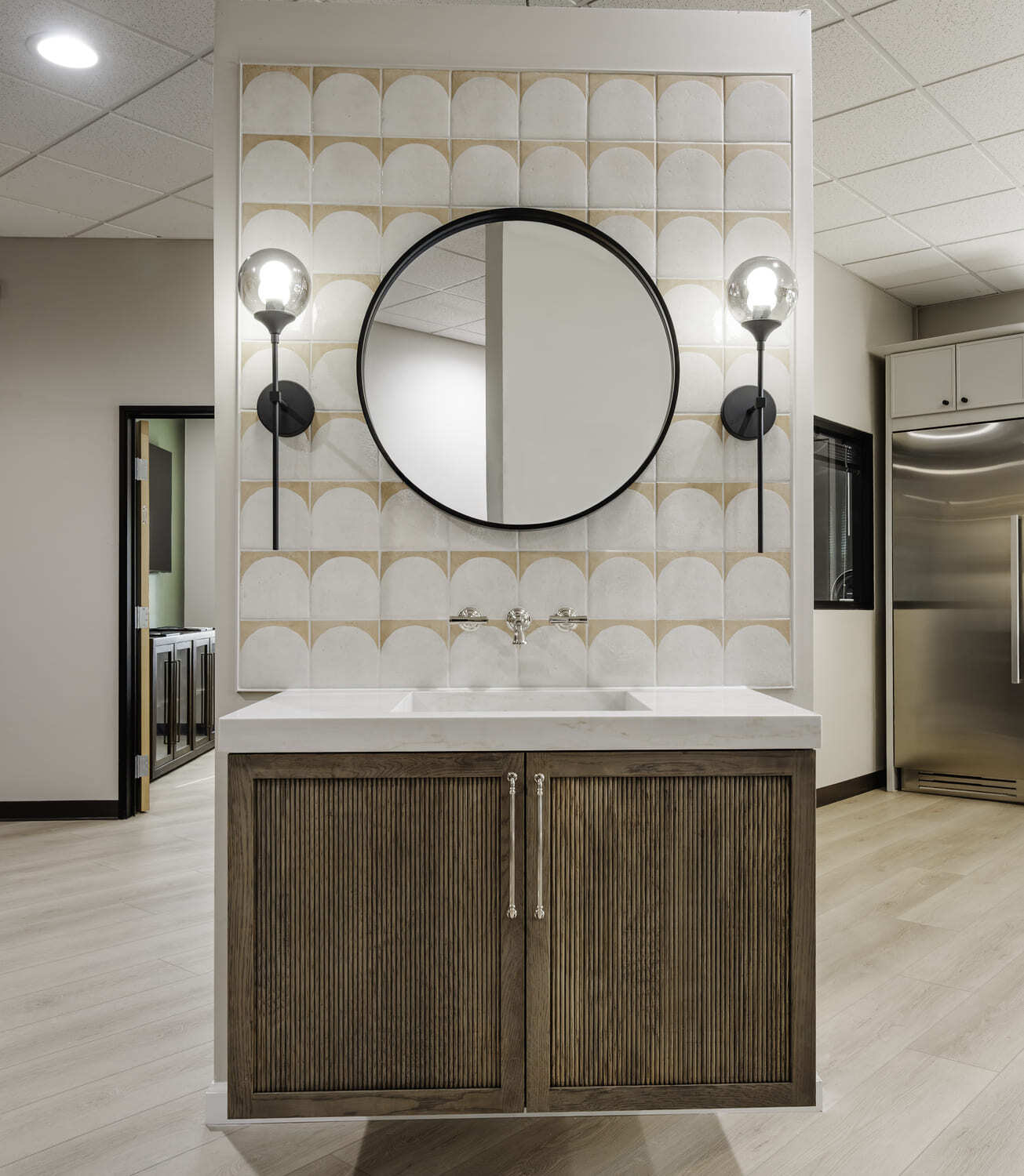 Bathroom showroom with round mirror, globe sconces, patterned tile wall, and wood vanity with white sink.