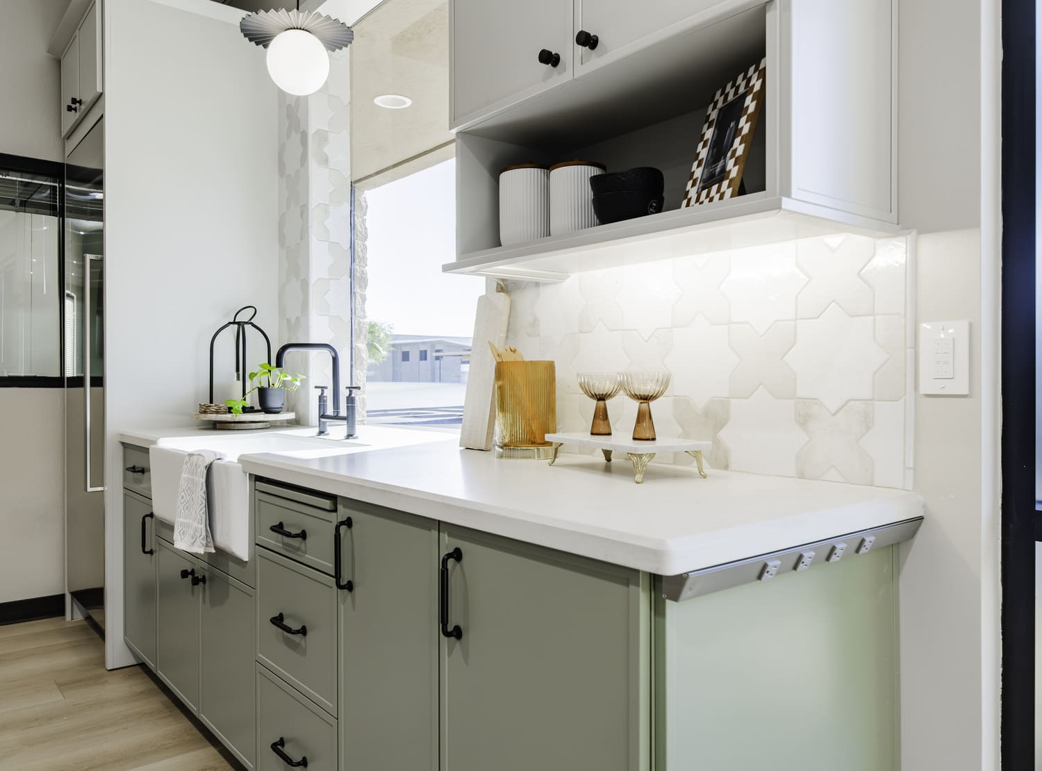 Kitchen showroom with sage green cabinets, white counters, open shelf, farmhouse sink, and patterned tile wall.