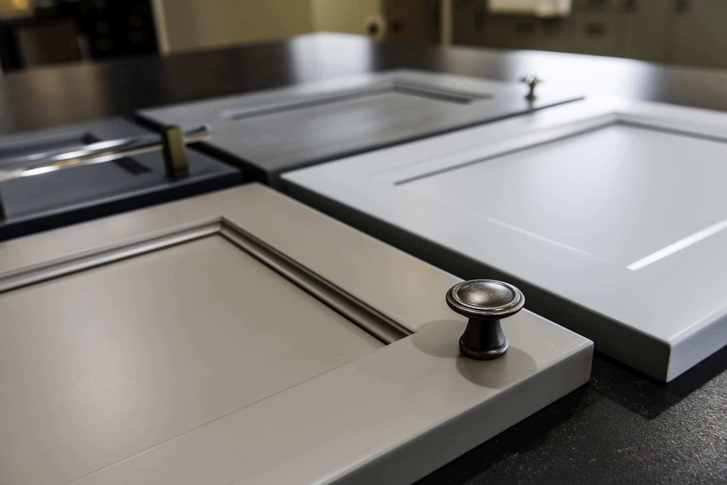 Taupe and white cabinet door samples with dark and metallic hardware on a dark kitchen showroom countertop.