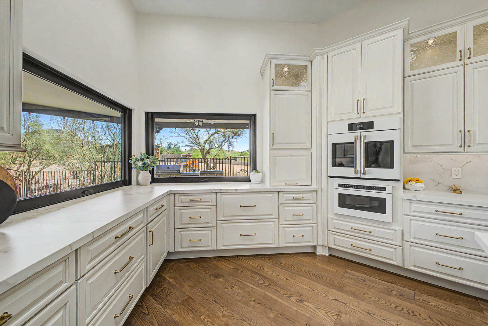 Bright kitchen remodel with white cabinets, large windows, built-in ovens, and warm wood flooring.